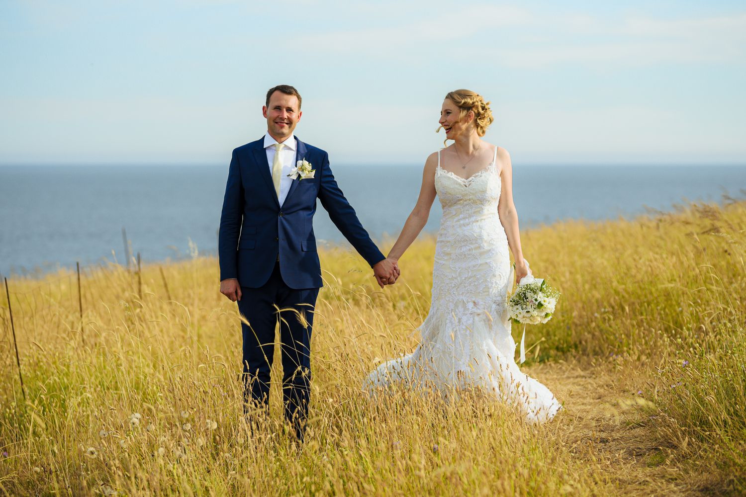 Bride and groom holding hands in golden field with ocean backdrop on their wedding day.