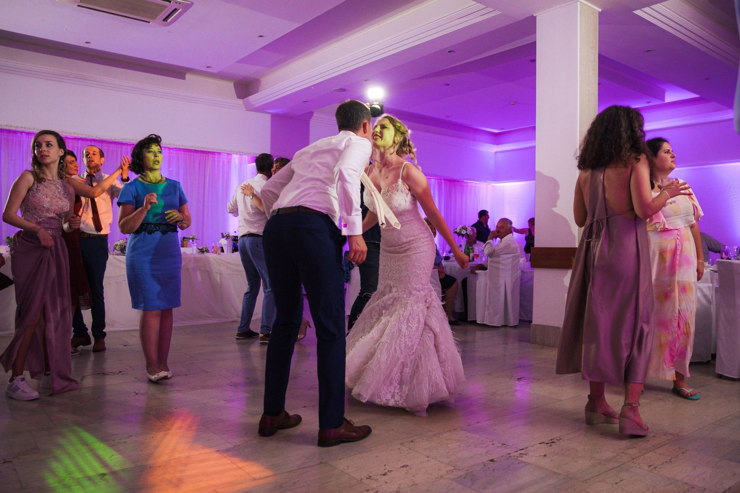 Bride and groom share a romantic dance surrounded by guests at purple-lit wedding reception.