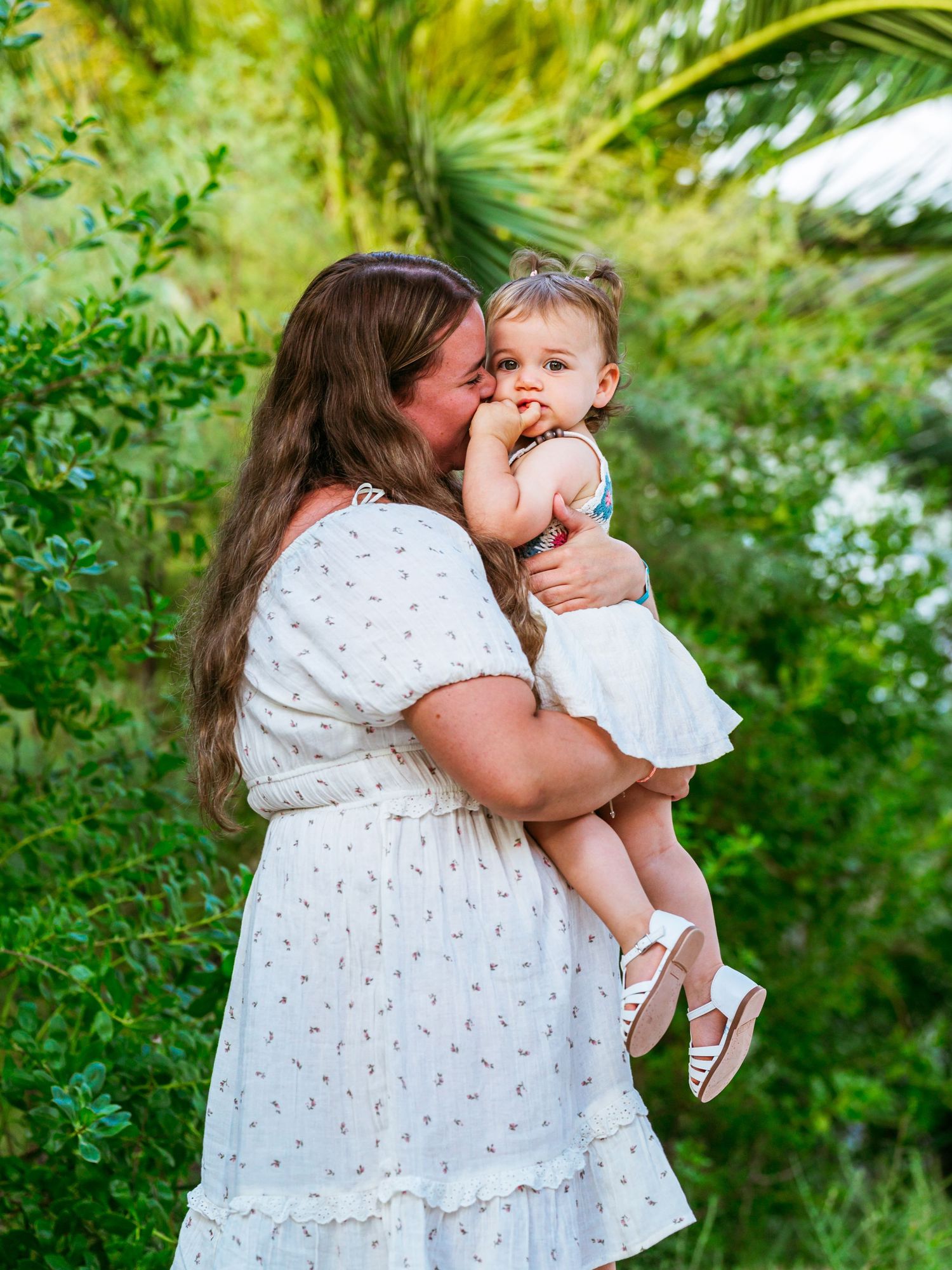A loving mother in a white floral dress kisses her baby girl outdoors surrounded by lush green foliage.