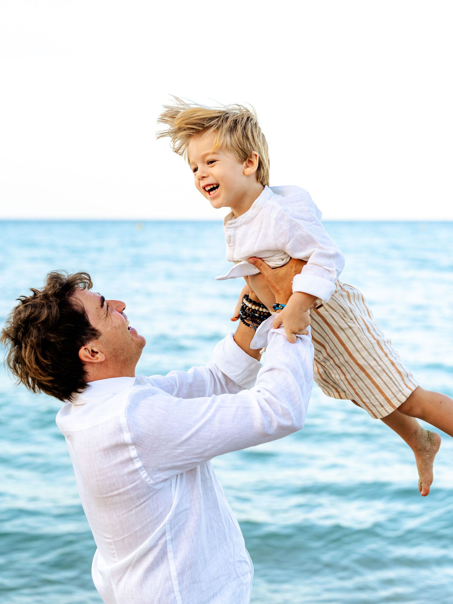 Father in white shirt lifting laughing blond toddler high at sunny beach.