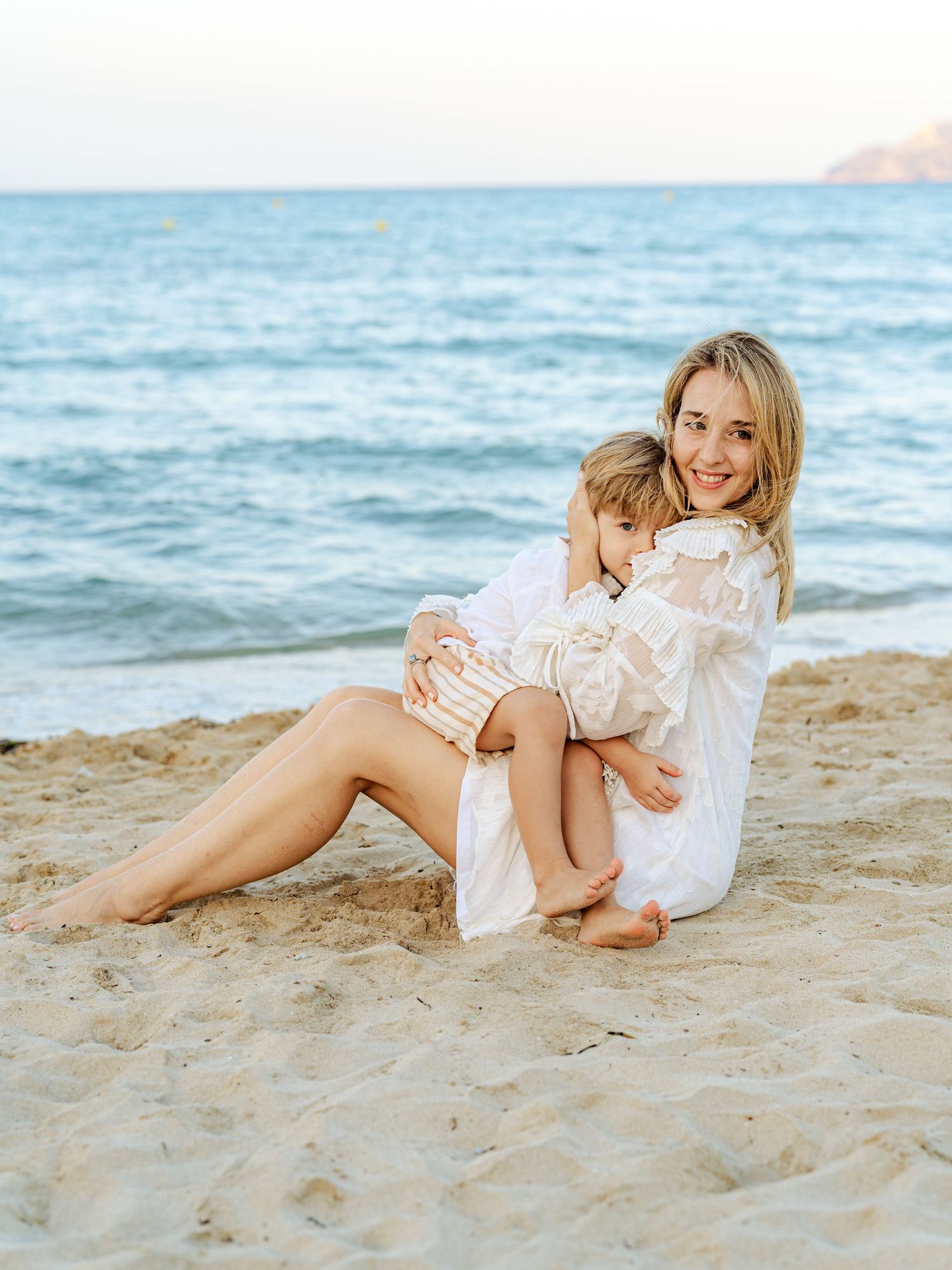 Smiling blonde mother in white dress hugging her young child while sitting on a sandy beach by the sea.