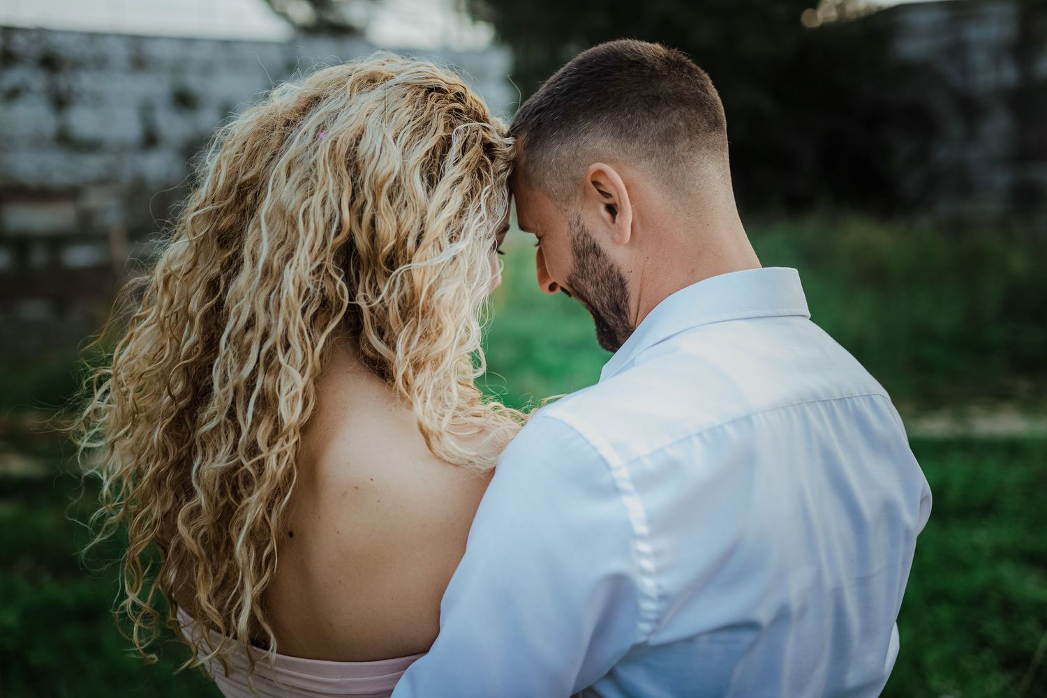 Romantic couple touching foreheads outdoors, woman with curly blonde hair in backless dress embracing man in white shirt.
