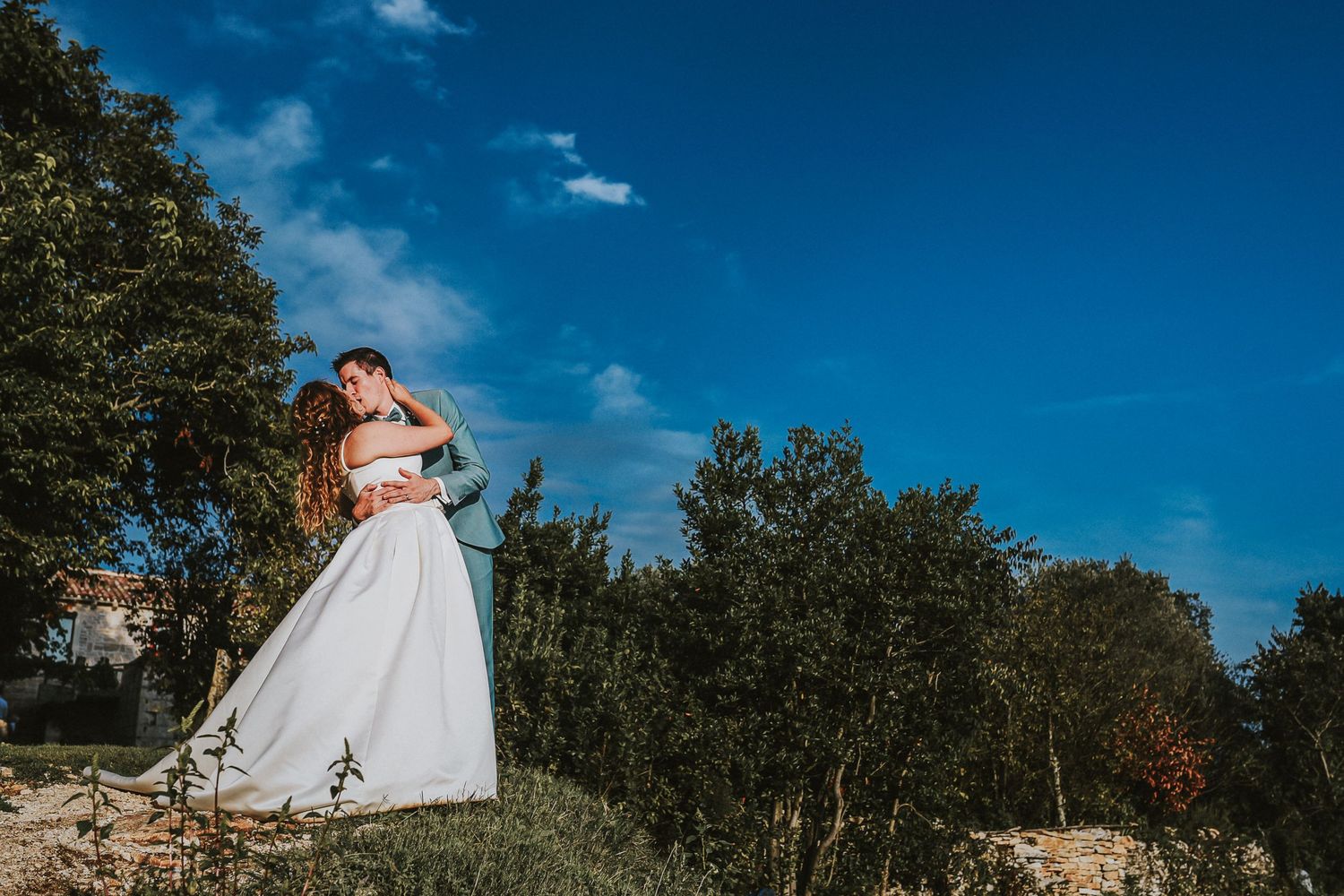 Bride and groom embracing against a vivid blue sky surrounded by green trees outdoors.