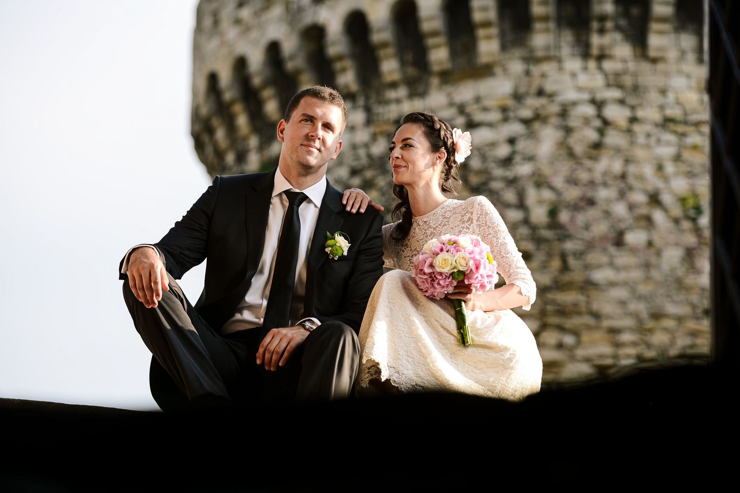 Wedding couple sitting together near a stone tower, bride holding a pink bouquet.