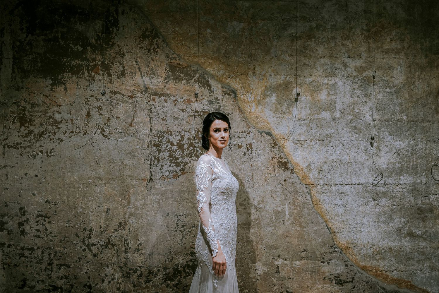 Elegant bride in a lace wedding gown posing gracefully against a rustic textured stone wall.