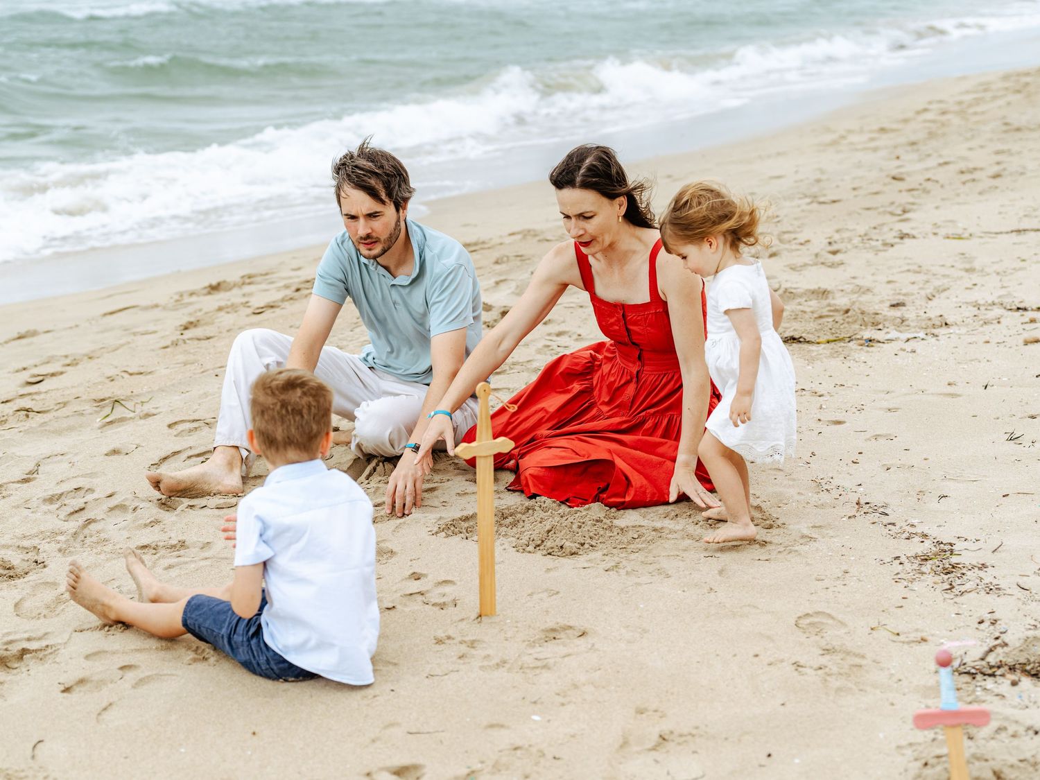 A family of four plays together on a sandy beach, with a mother in a red dress and father with two children.