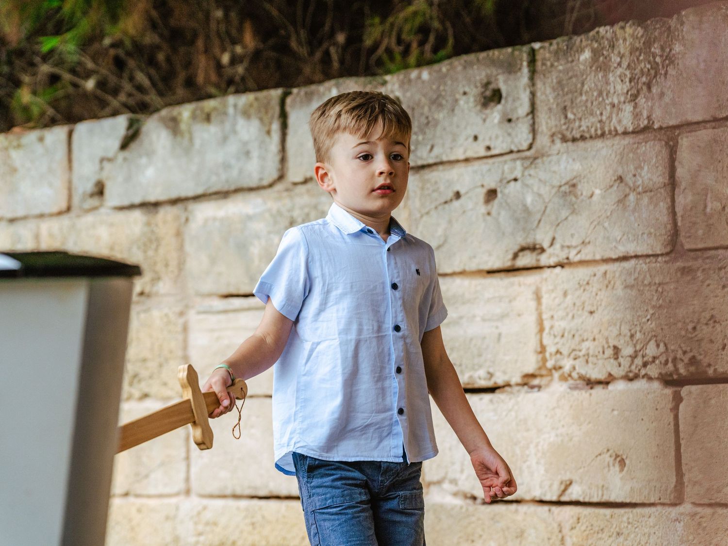 Young blond boy in light blue shirt holding wooden toy sword near stone wall outdoors.