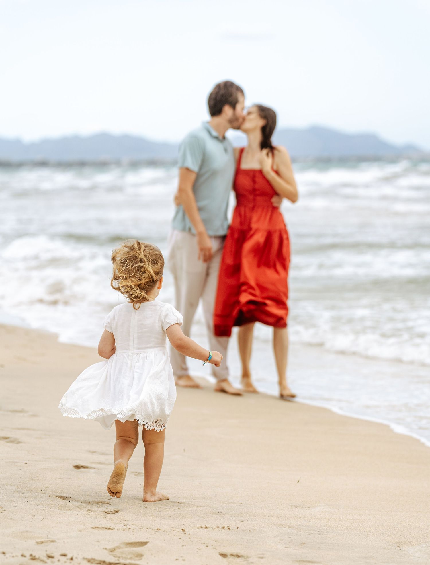 Young child in white dress running toward parents kissing on a sandy beach with mountains behind.