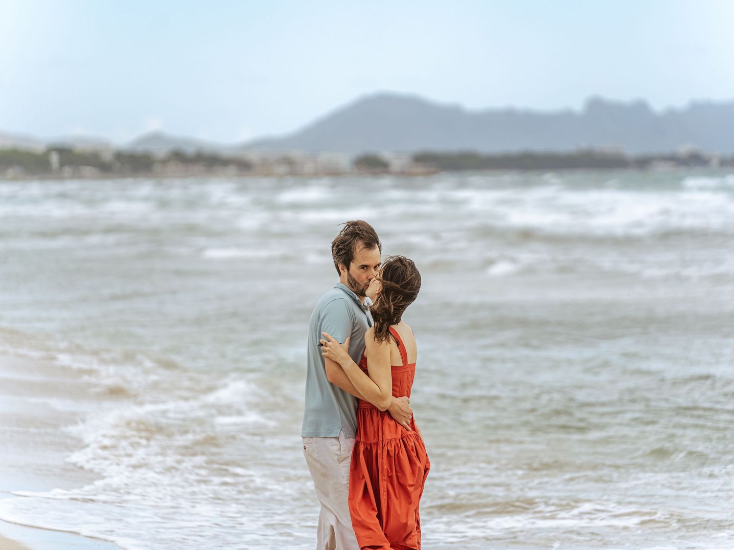 Couple sharing a tender moment on a windy beach, woman in red dress, mountains in background.