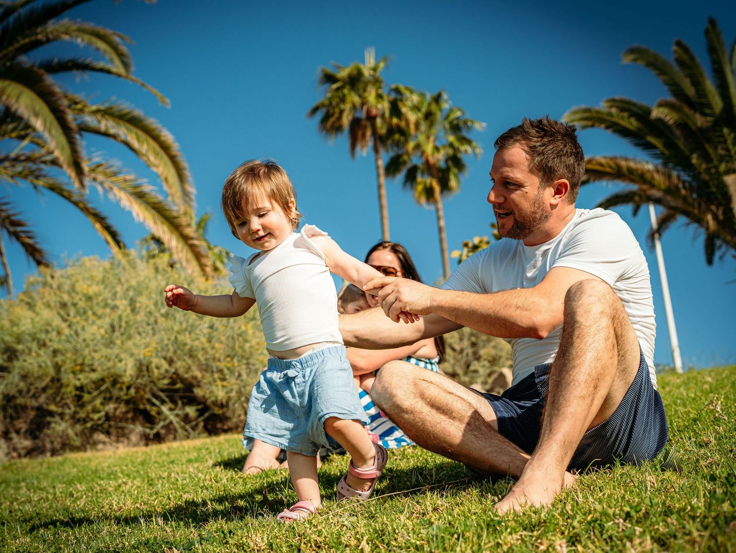Parents helping excited toddler walk on green grass under blue sky with palm trees.