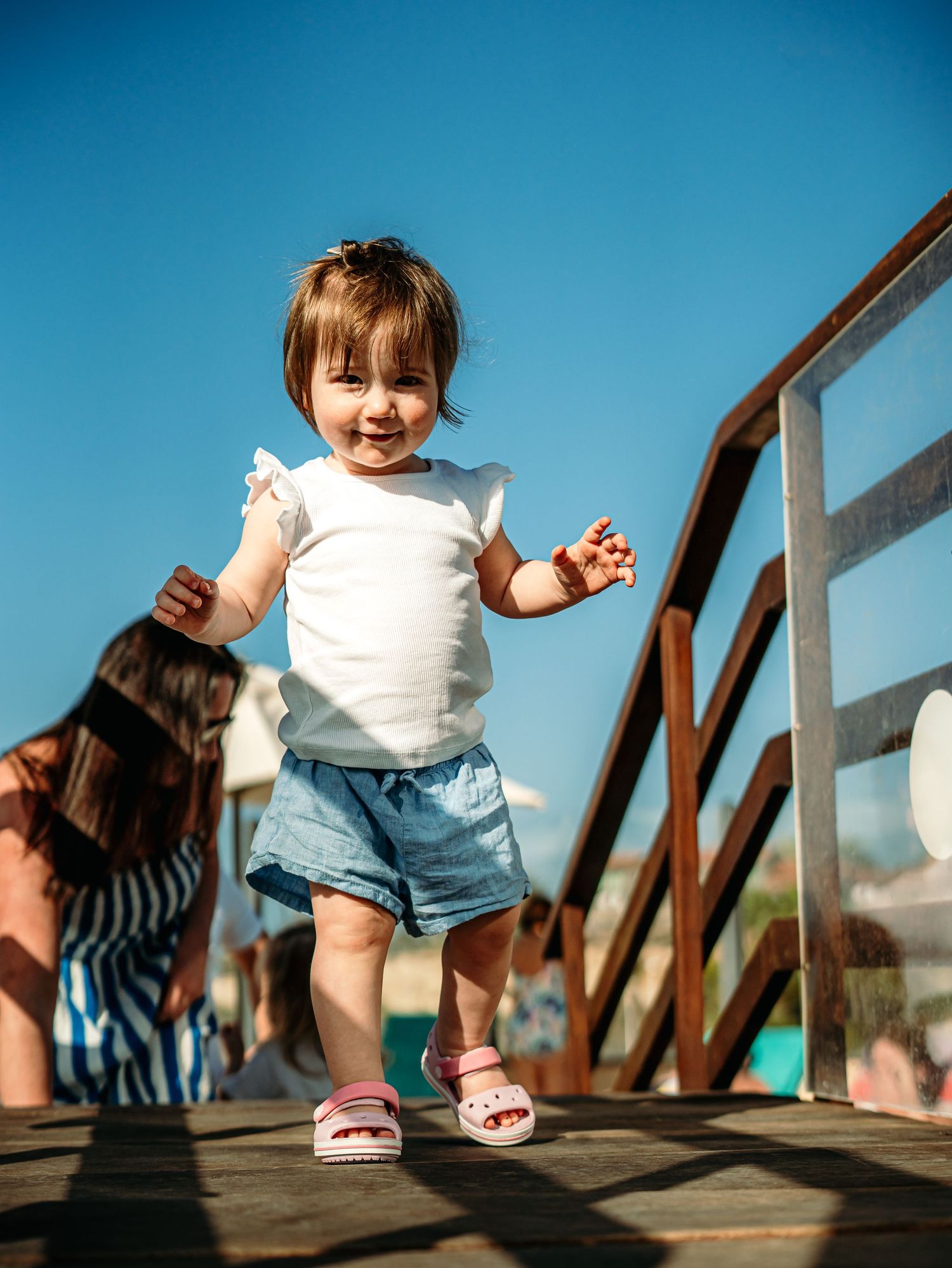 Smiling toddler girl in white top and denim shorts walking on a wooden boardwalk under blue sky.