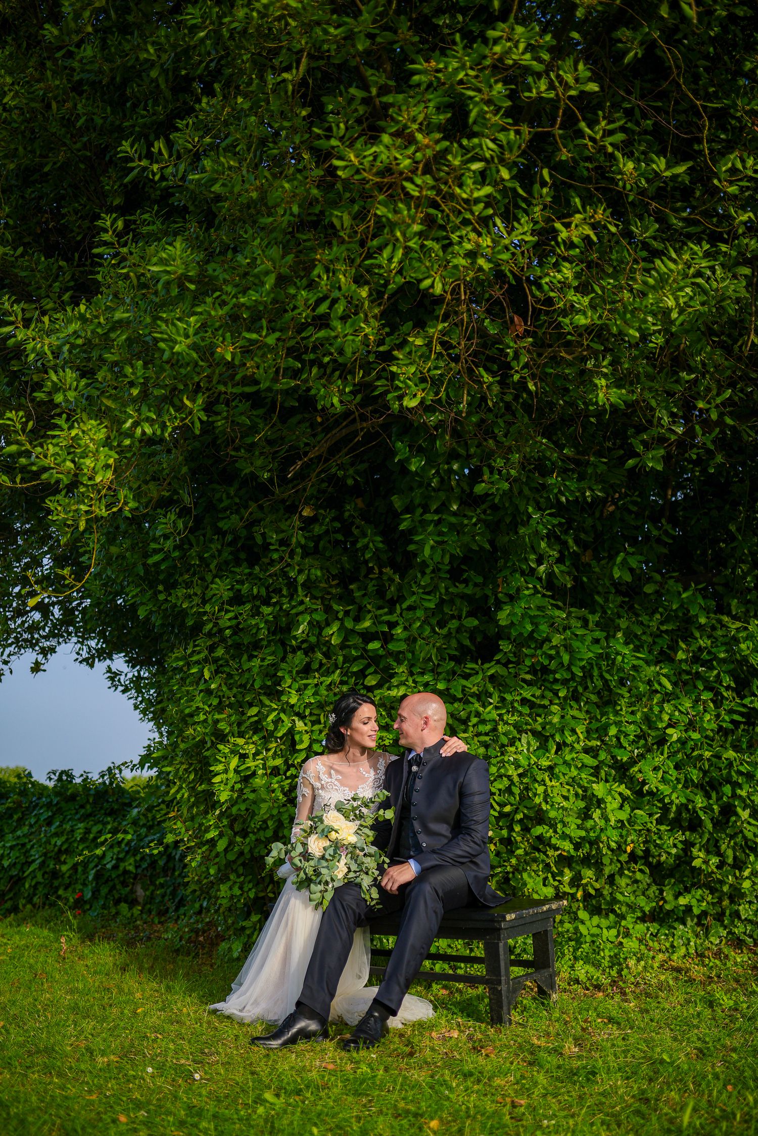 Bride and groom sitting together on a bench surrounded by lush green trees at sunset.