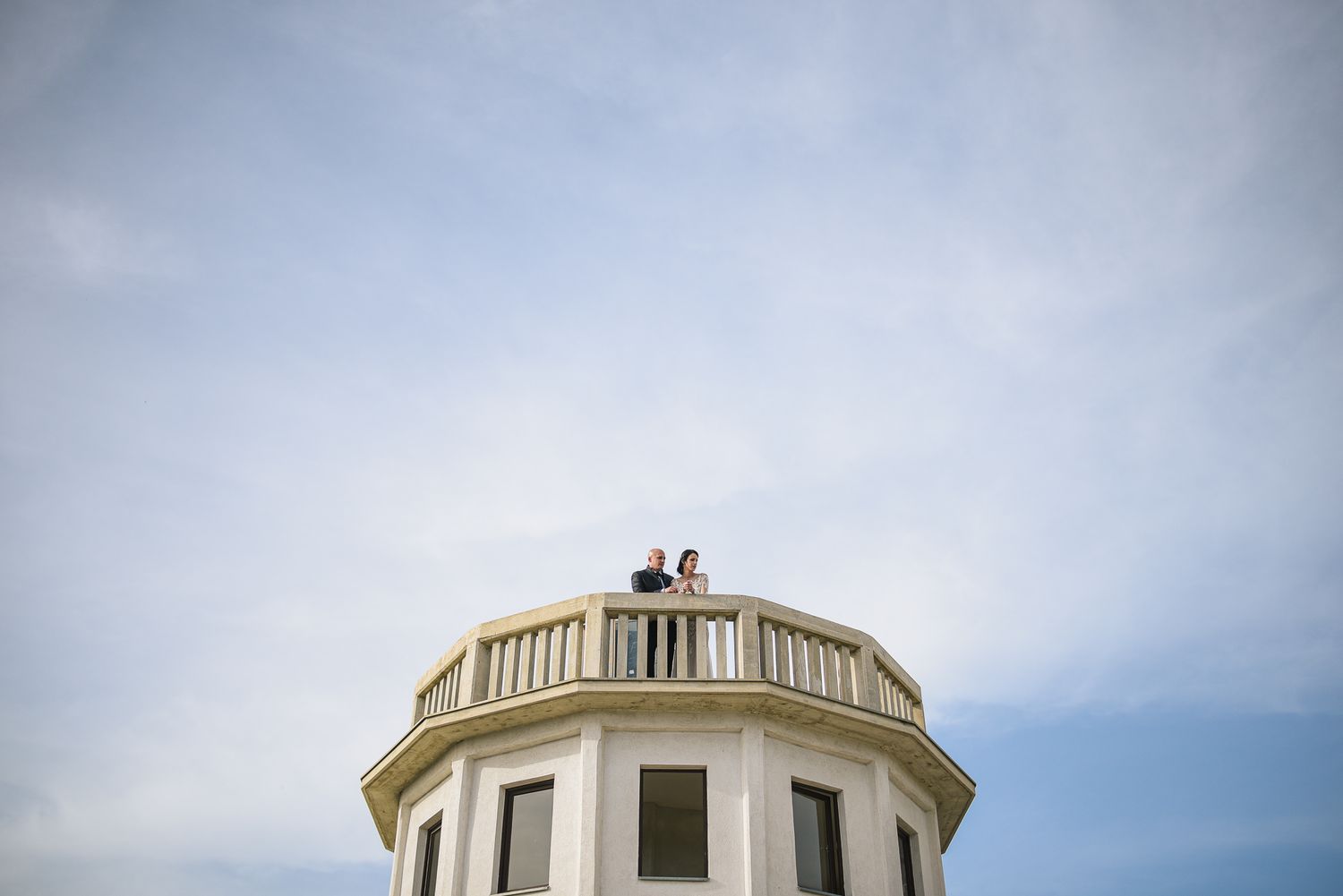 Bride and groom stand together on a circular tower balcony against a bright blue sky.