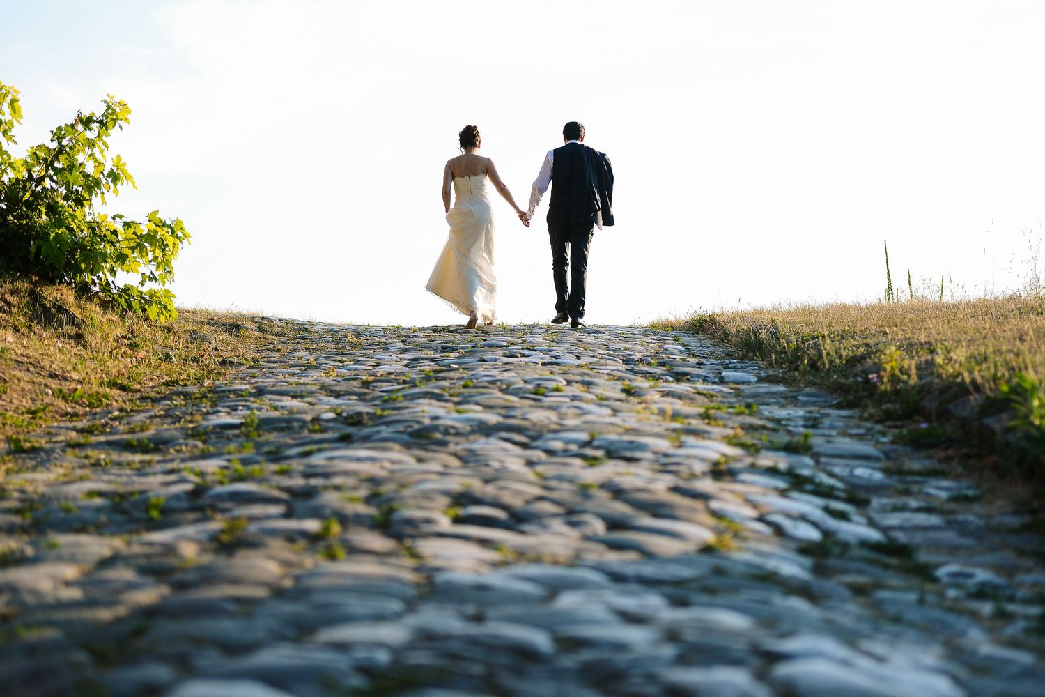 Bride and groom walking hand in hand along a cobblestone path during their outdoor wedding portrait session. By Herzenshaus Studio.