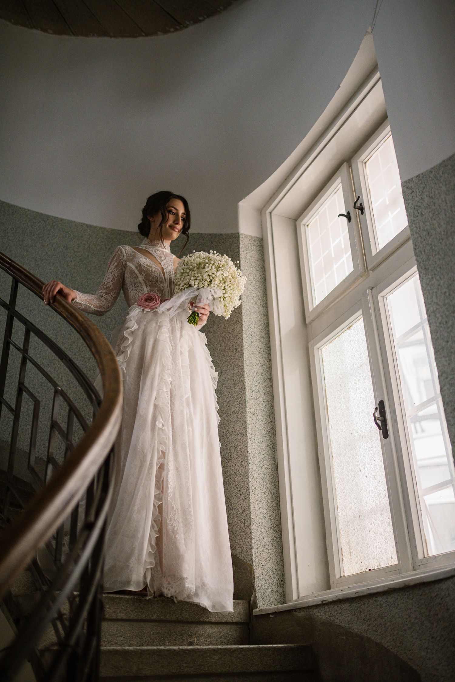 Elegant bride in pink lace gown holding white bouquet on spiral staircase.