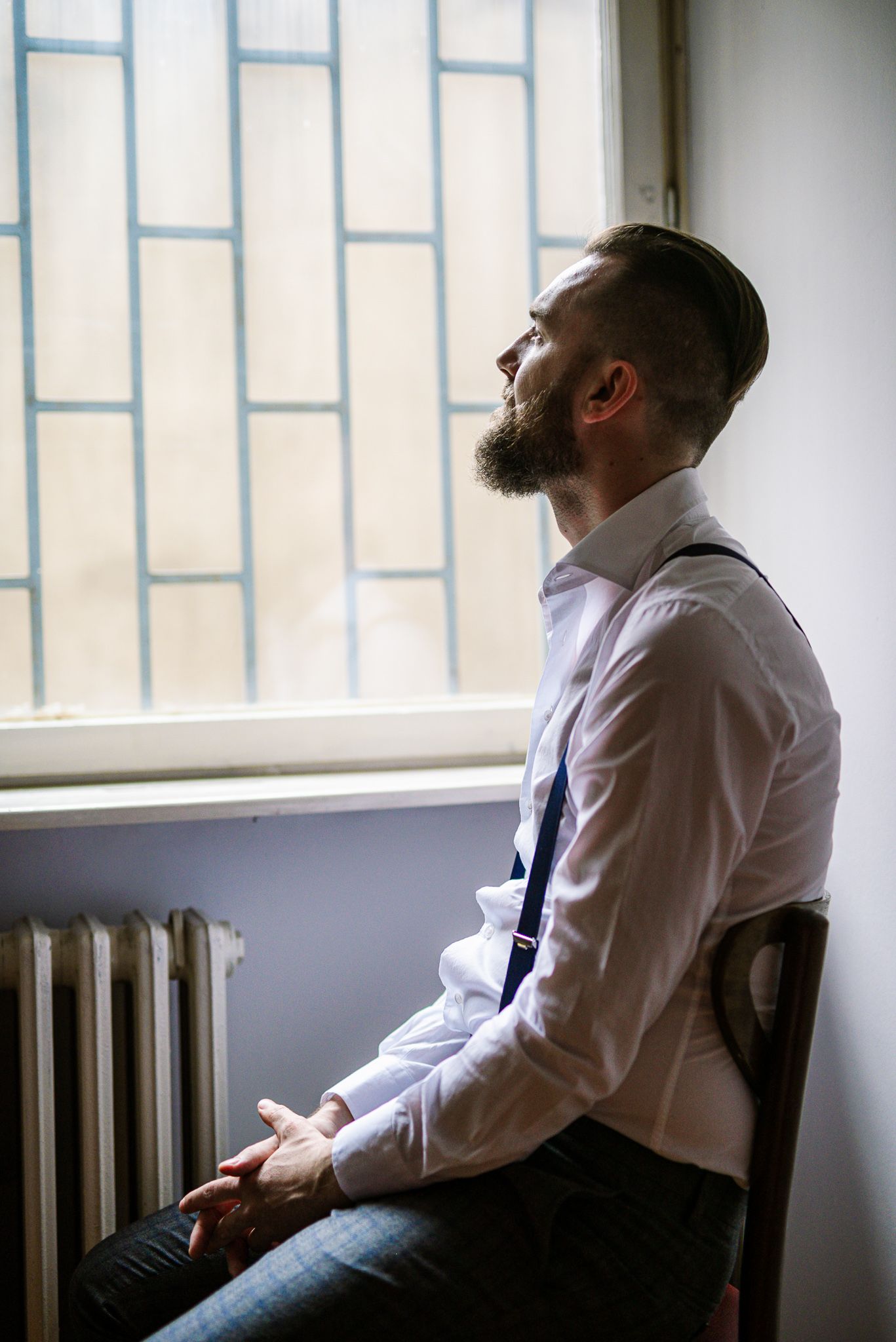 Bearded groom in white shirt and suspenders sitting by a window in contemplative moment before wedding.