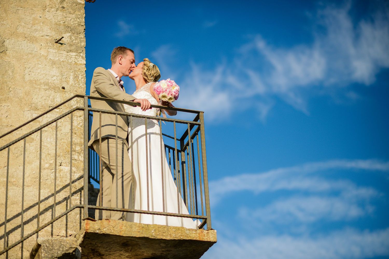 Wedding couple kissing on a rustic stone balcony against a vivid blue sky.
