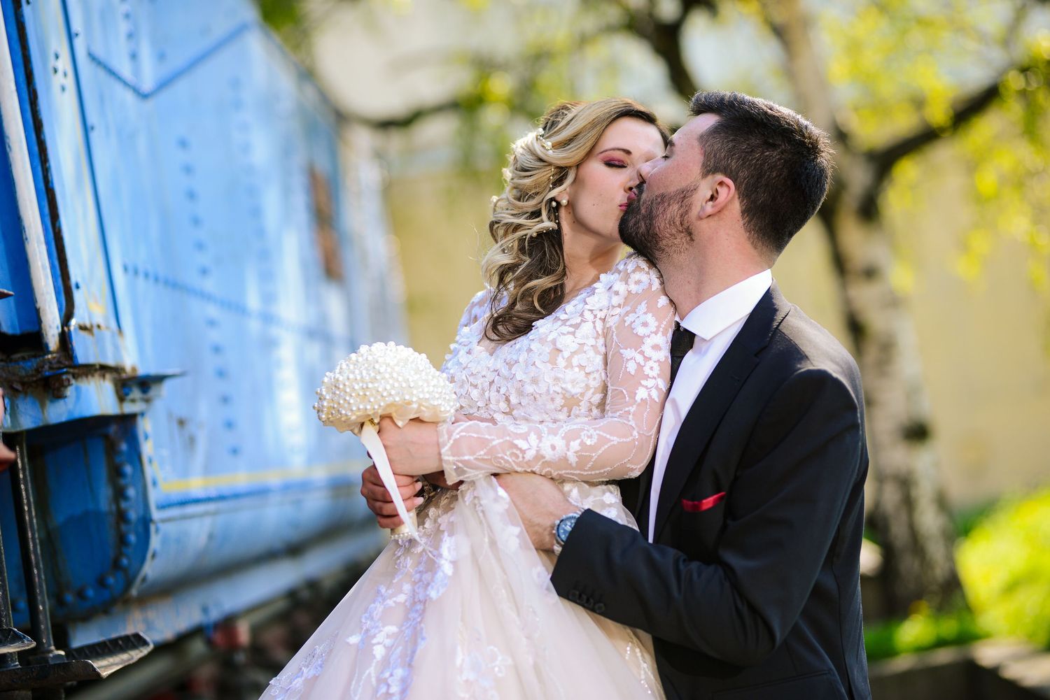 Groom kisses bride on the cheek as she holds a white bouquet near a vintage blue train car.