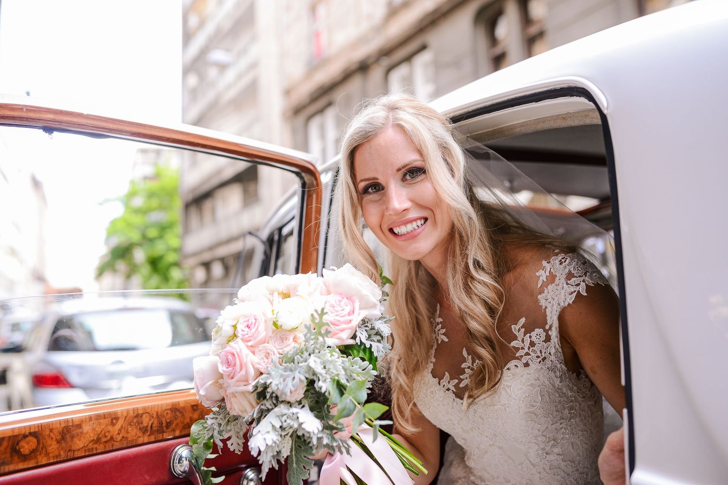 Smiling blonde bride holding a pink and white bouquet stepping out of a vintage wedding car.