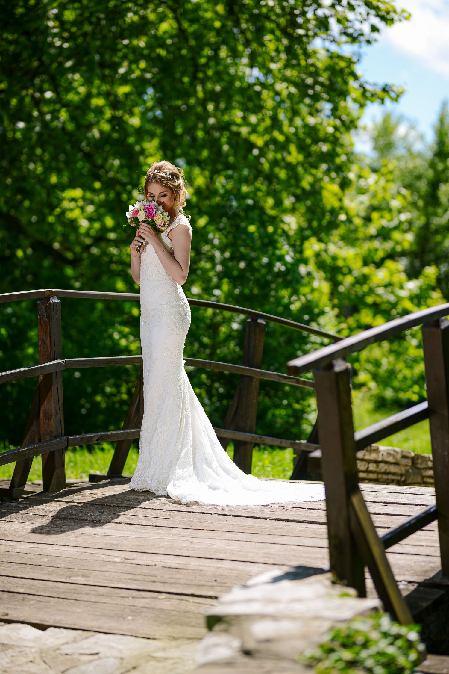 Bride in a fitted white lace gown smells her bouquet while standing on a wooden bridge in a sunny garden.