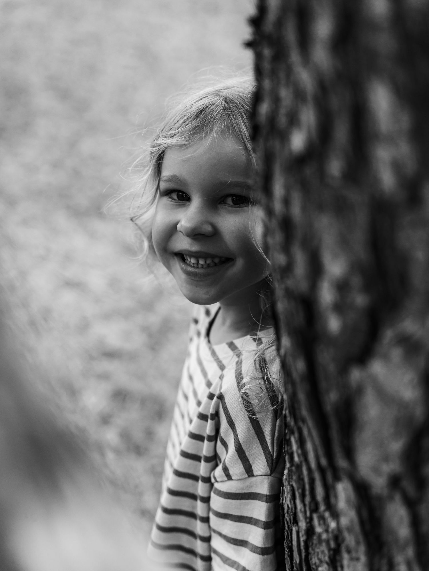 Black and white portrait of a cheerful blonde girl peeking and smiling from behind a tree trunk.
