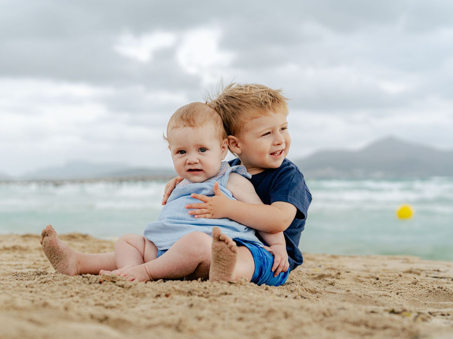 Two young siblings hugging and smiling together on sandy beach with ocean backdrop.