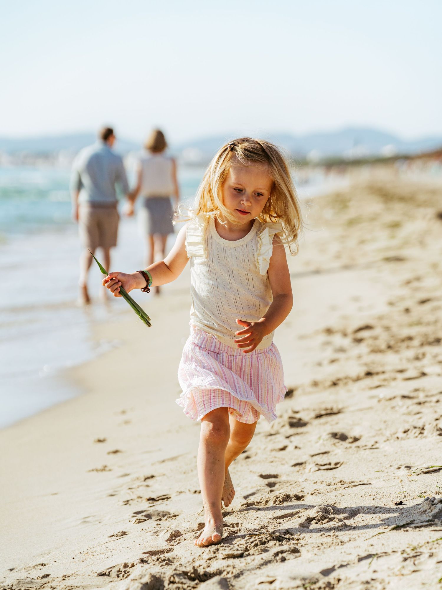 Young blonde girl running barefoot on a sandy beach while parents walk hand in hand behind her.