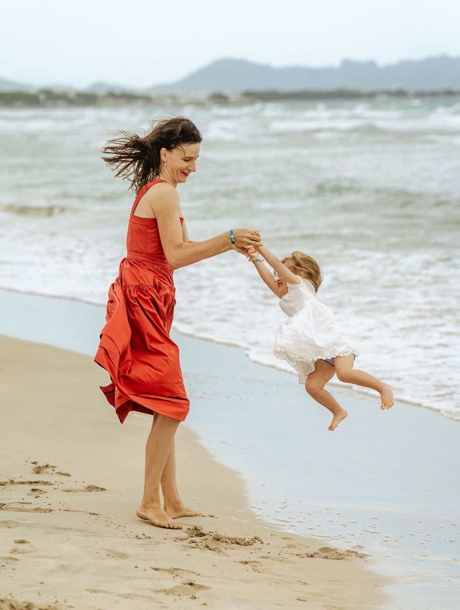 Joyful mother in red dress swinging young child by the hands on a beach.