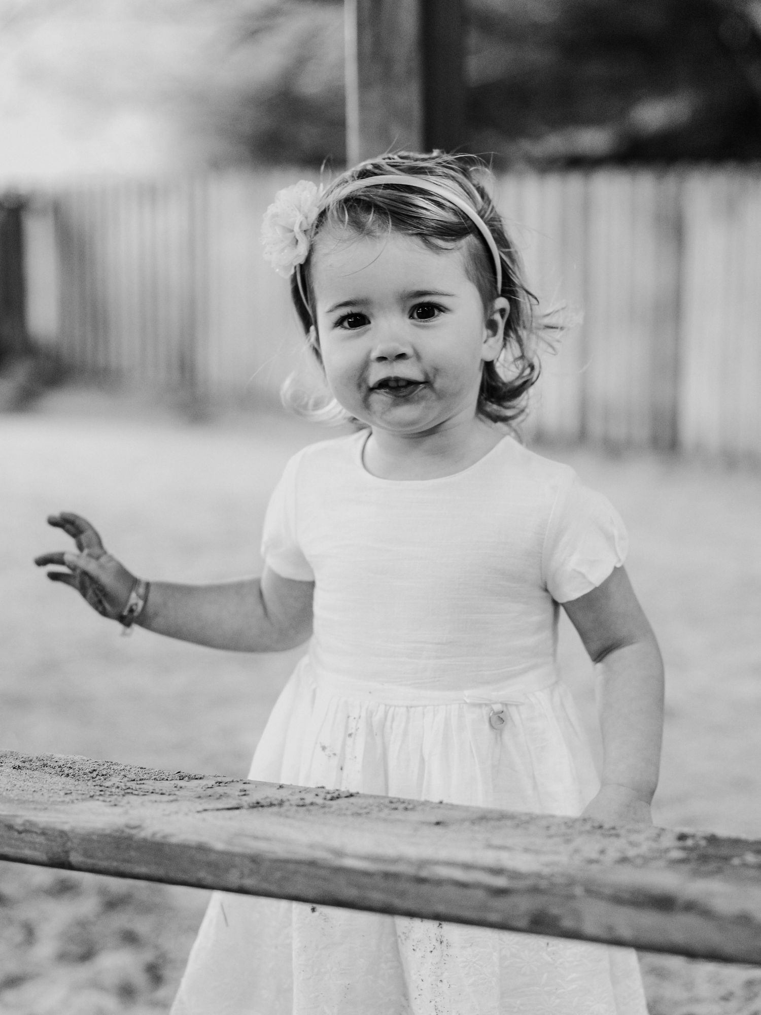 Black and white portrait of smiling toddler girl in white dress at outdoor playground.