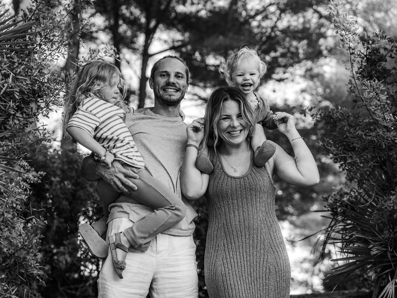 Black and white photo of a joyful family of four among trees, parents holding two young children.