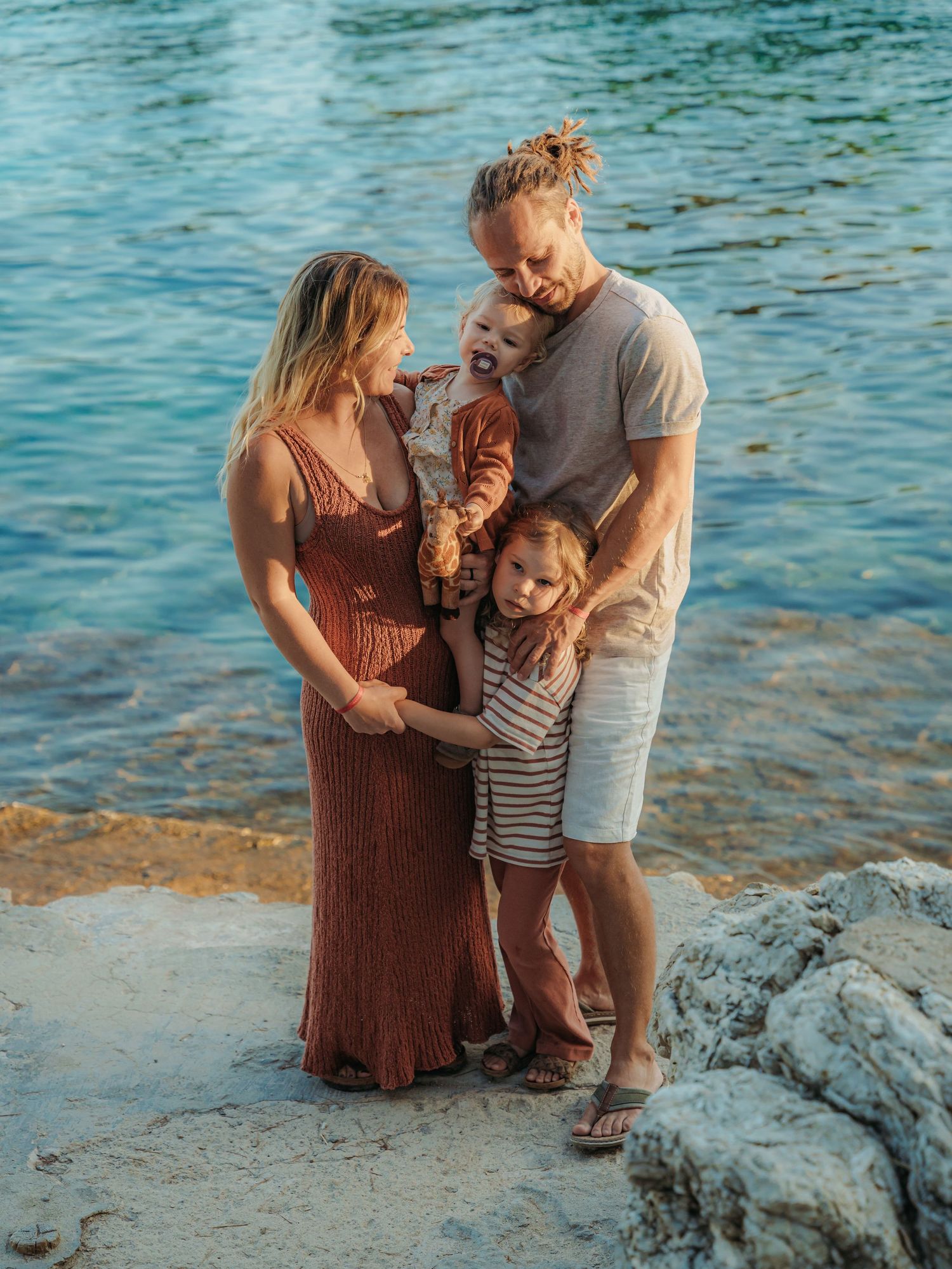 Family of four poses by the sea, pregnant mother in rust dress with father holding toddler and young child beside them.