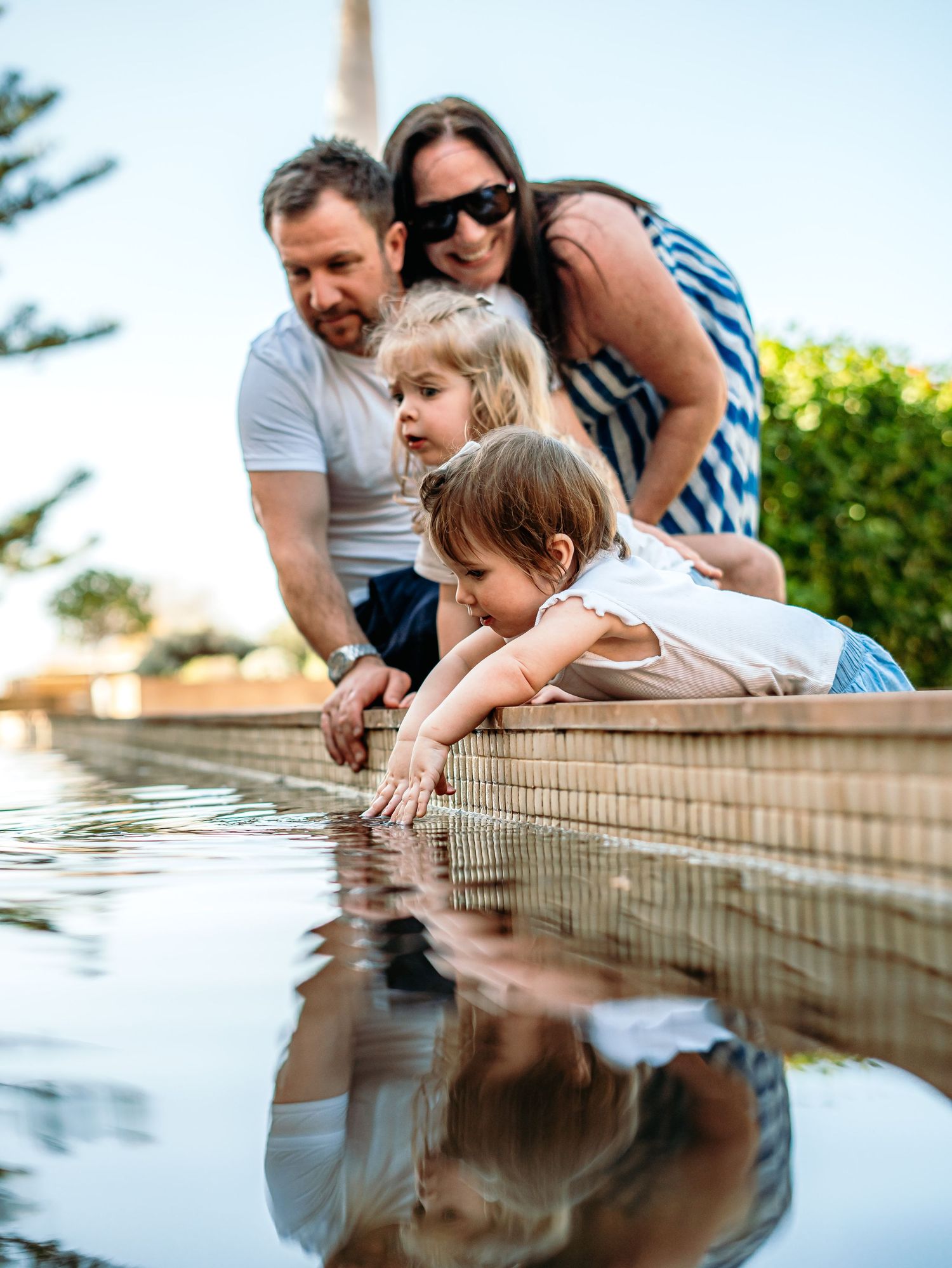 Parents and two young daughters explore a calm reflective garden water feature together outdoors.