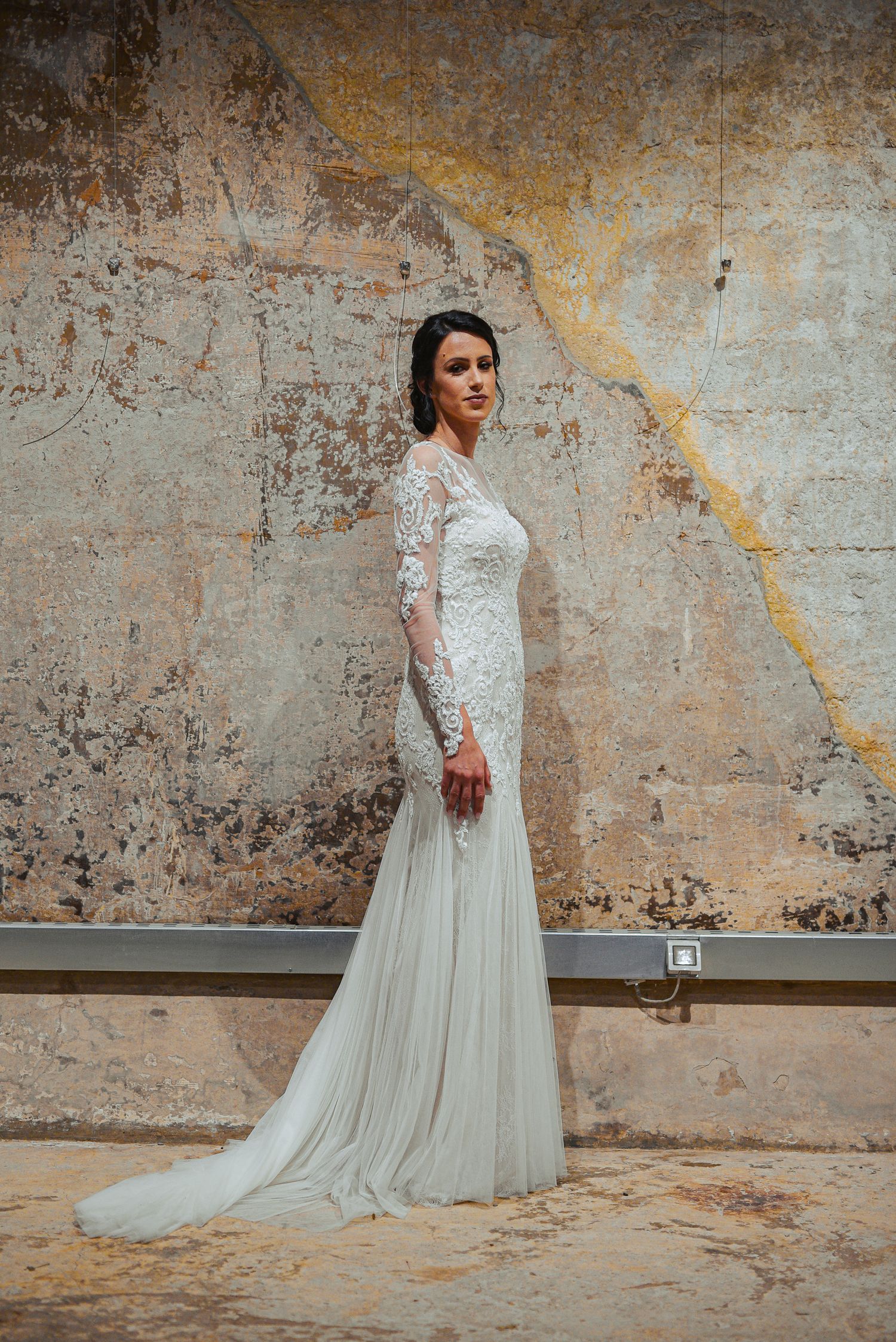 Bride in a fitted long-sleeve lace gown with a flowing train poses by a rustic wall.