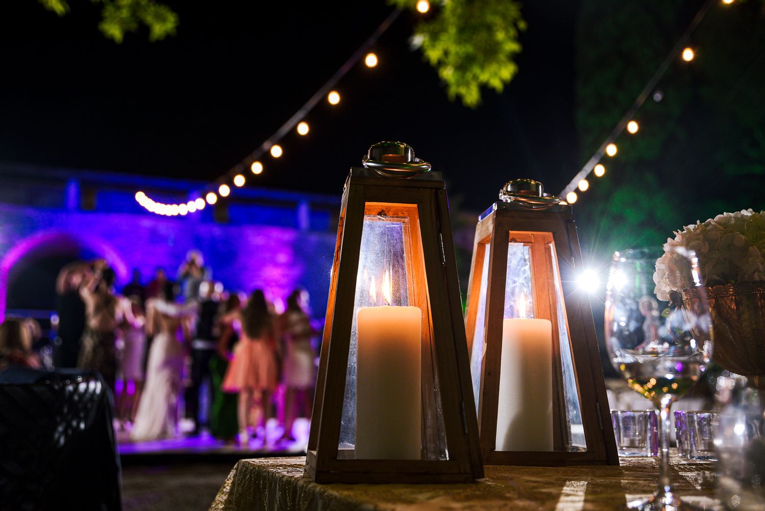 Decorative candle lanterns on a wedding table with guests dancing in the background at night.