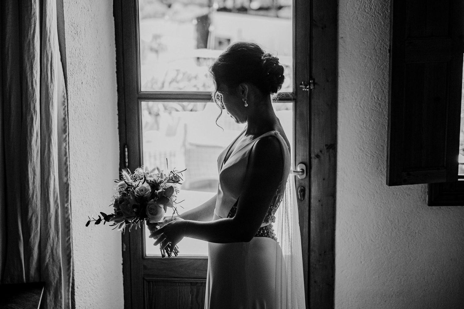Black and white portrait of bride in wedding dress holding bouquet by open window.