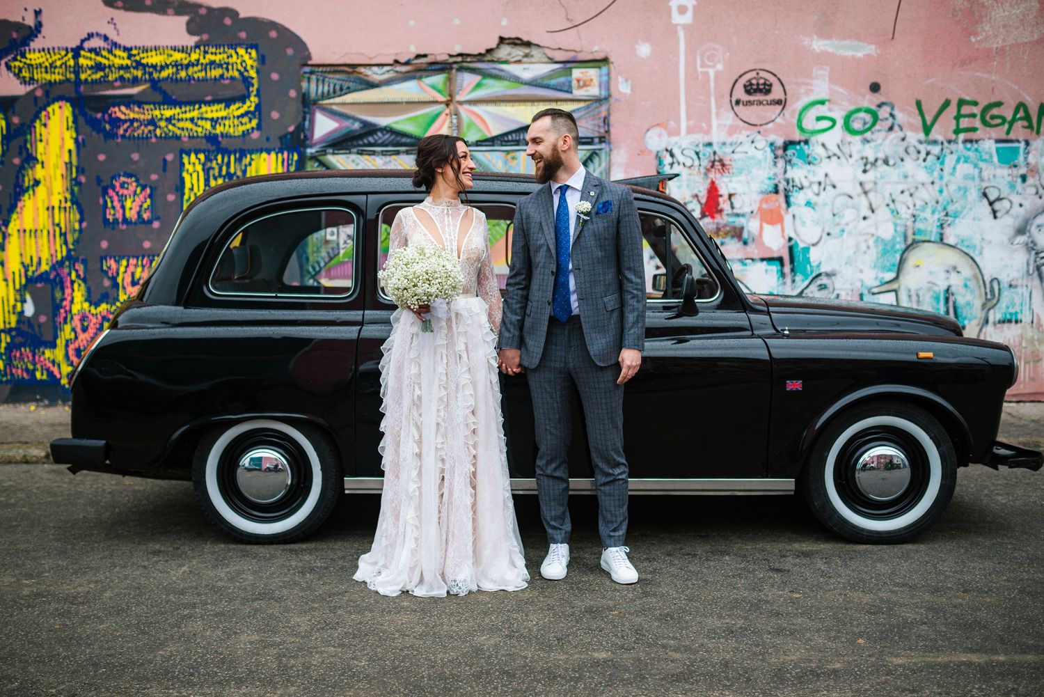 Bride and groom standing before vintage black London taxi with vibrant graffiti mural on wedding day.