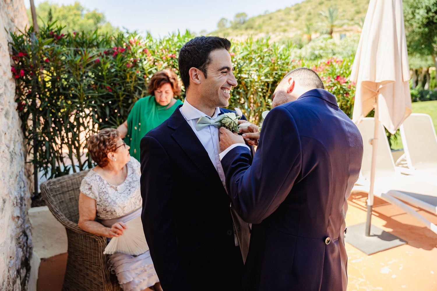 Groom in navy suit having his tie adjusted by a guest at an outdoor wedding venue.