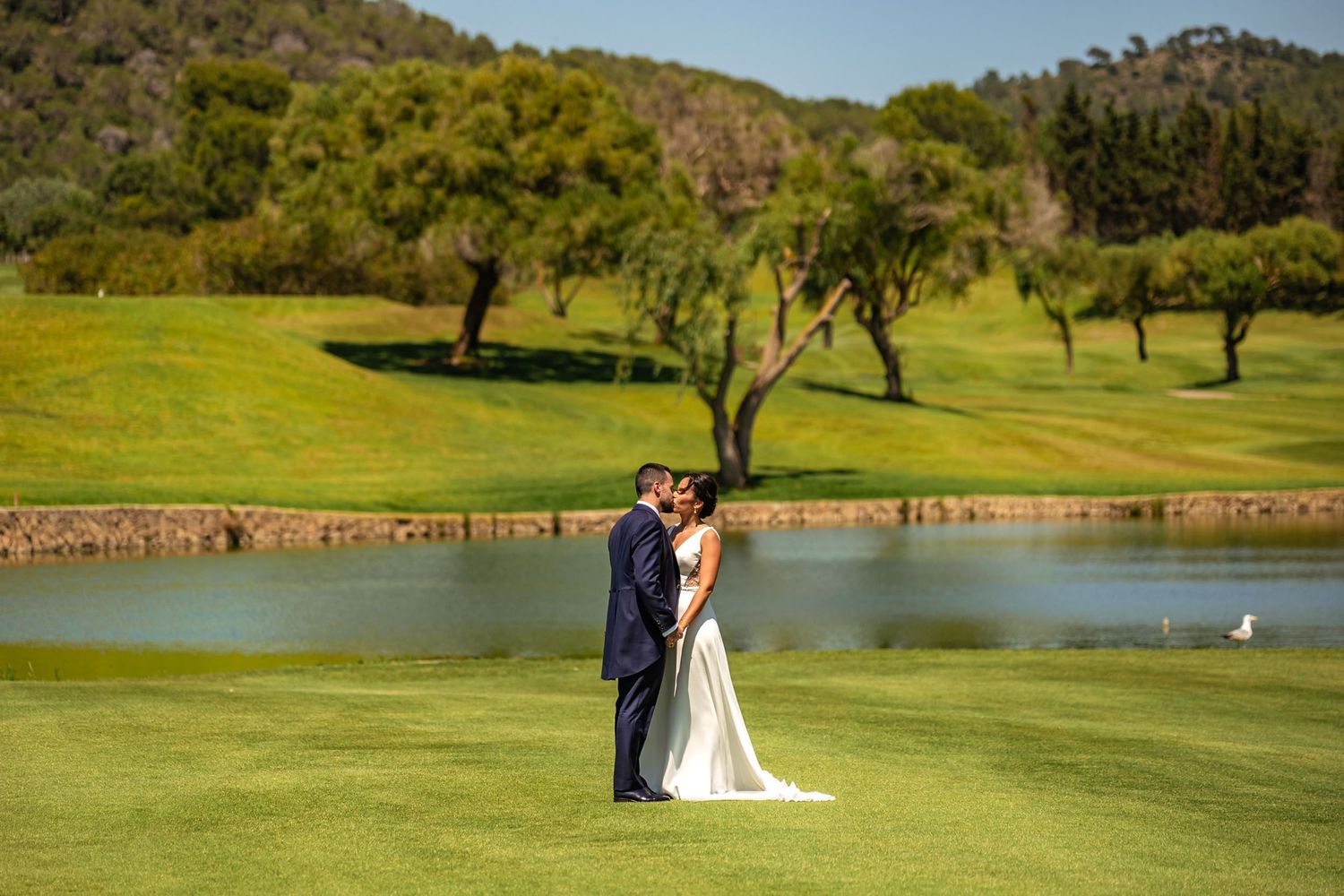 Bride and groom sharing a romantic kiss beside a tranquil pond on a lush green golf course.