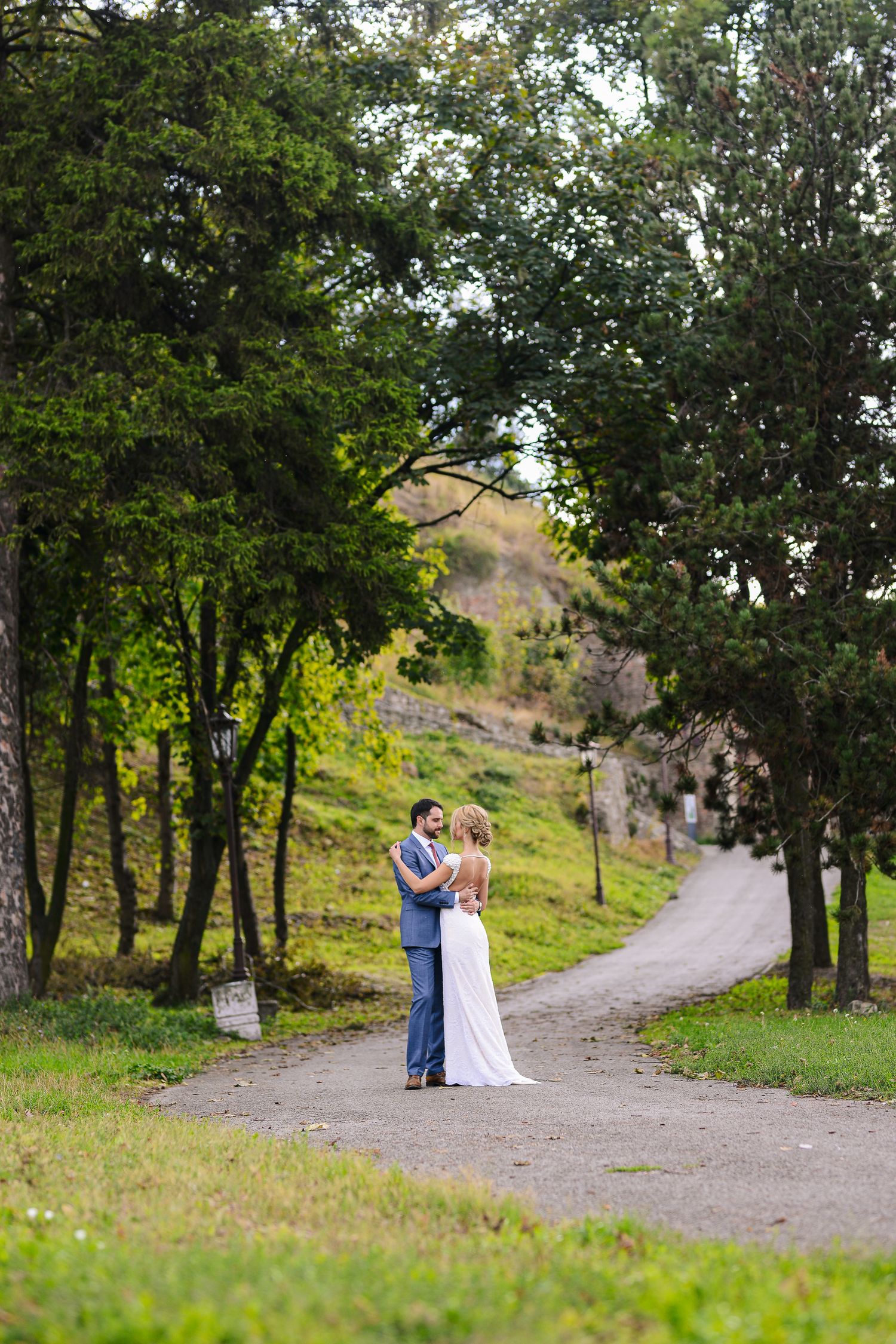 Bride and groom share a romantic embrace on a winding tree-lined path surrounded by lush green scenery.