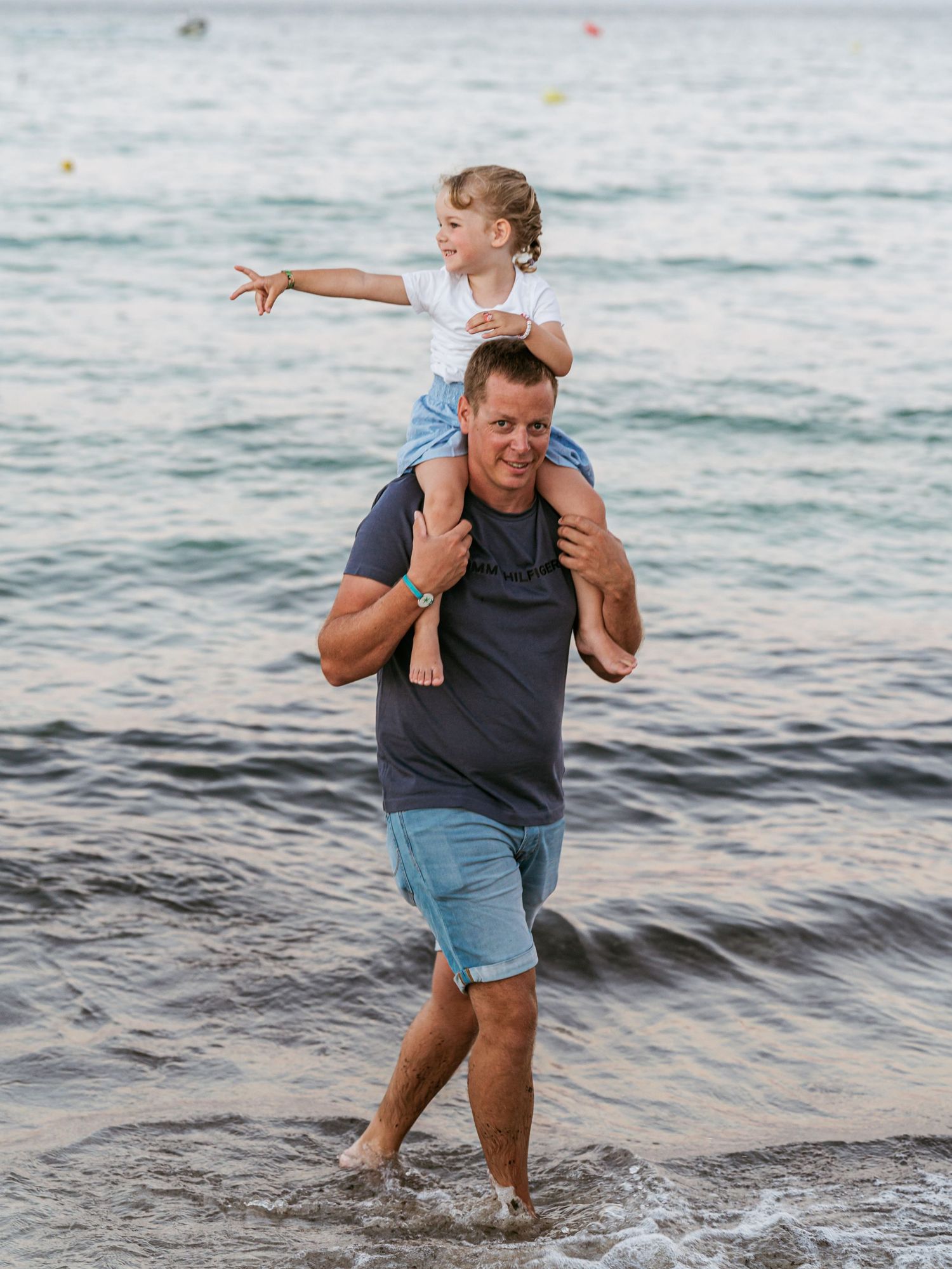 A father in a navy shirt carrying his young daughter on his shoulders while wading in the ocean.