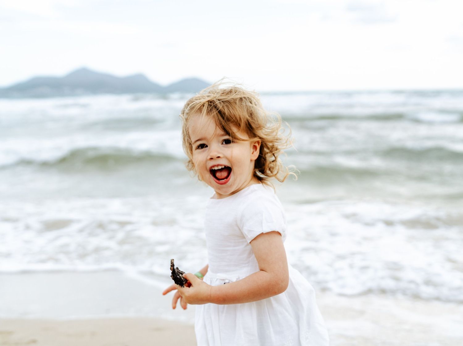 Joyful toddler girl in white dress laughing on beach.