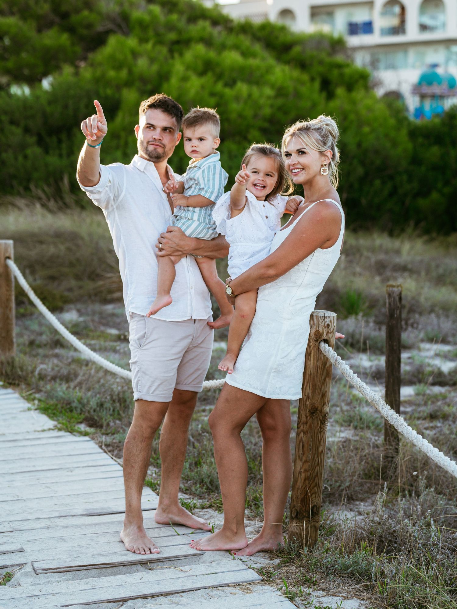 Young family of four poses on wooden boardwalk near beach during warm golden sunset.