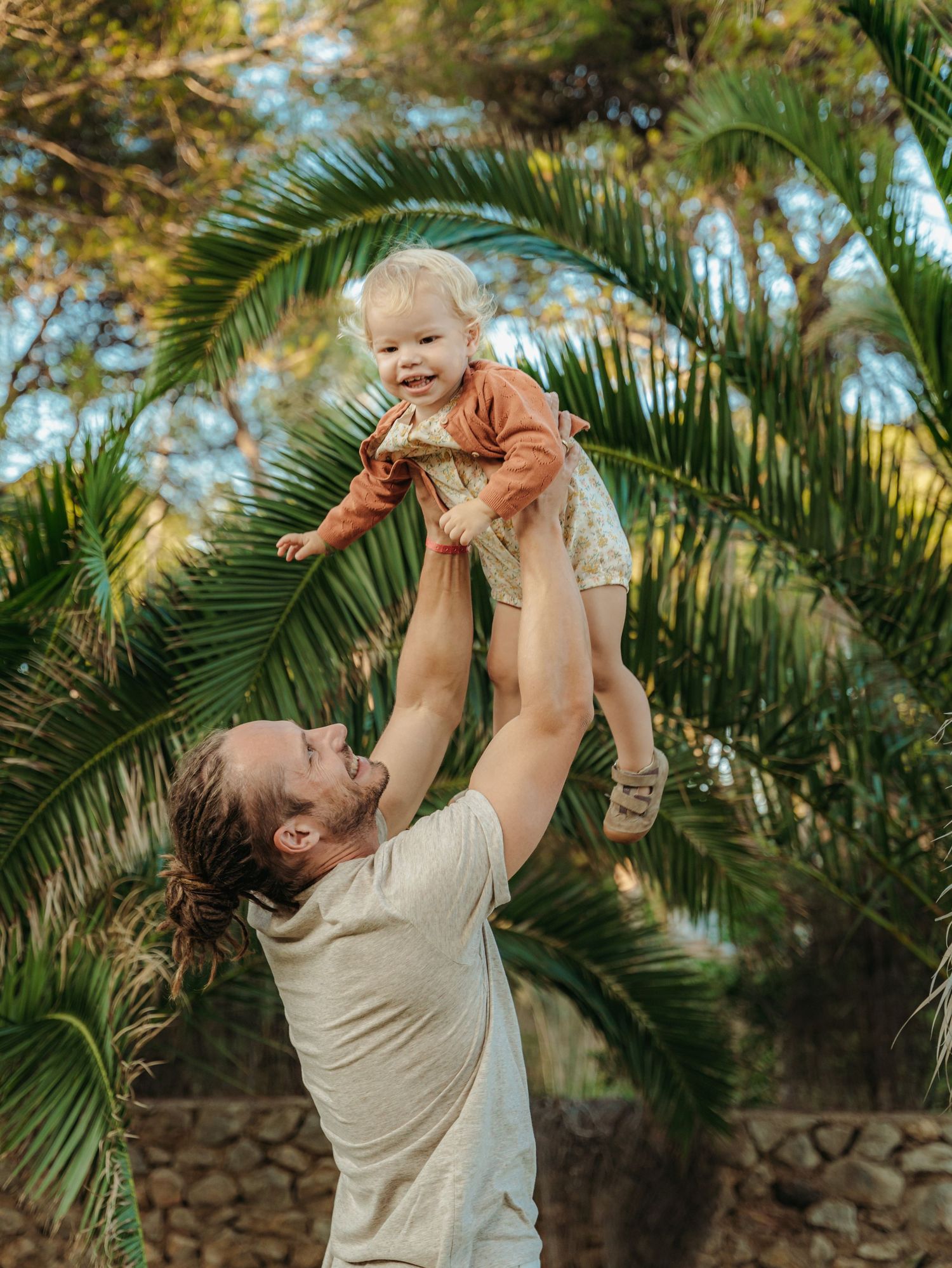 Father lifting happy laughing baby into air with tropical palm trees in background.