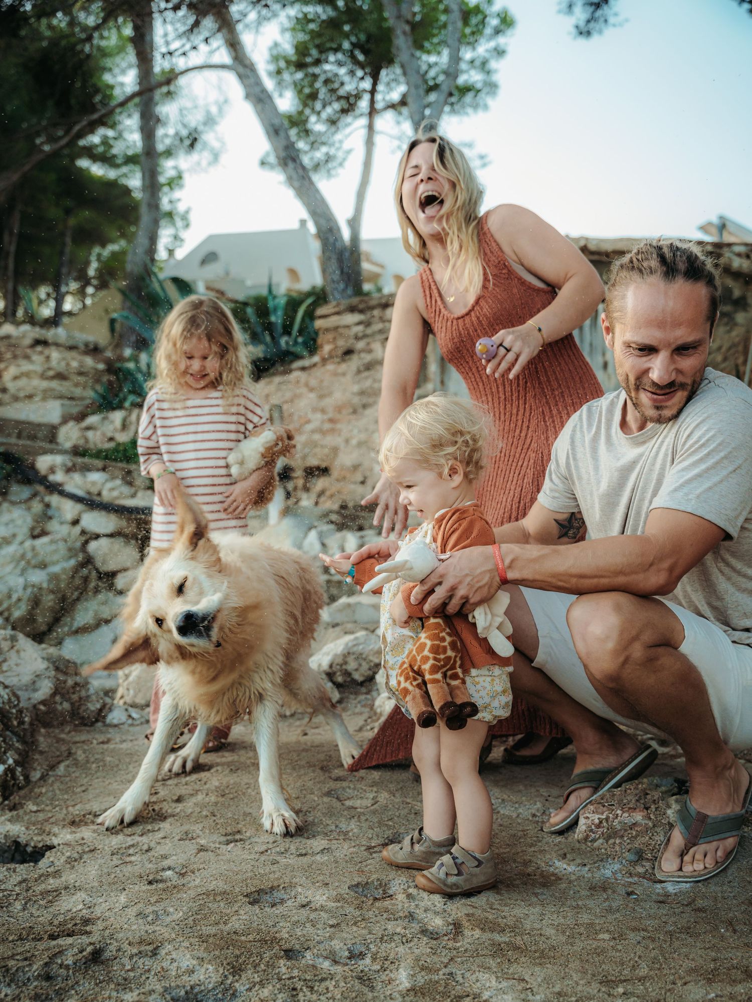 A laughing family enjoys a playful moment with a wet dog on a rocky outdoor landscape.