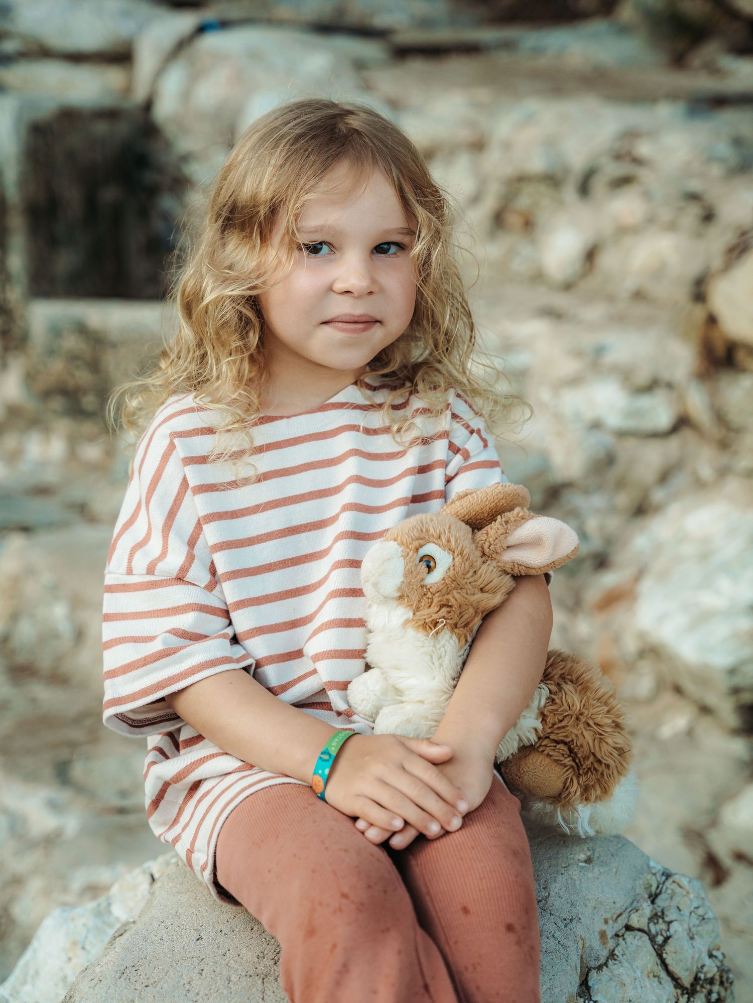 Blonde curly-haired girl in striped shirt hugging stuffed bunny while sitting on stone in rustic outdoor setting.