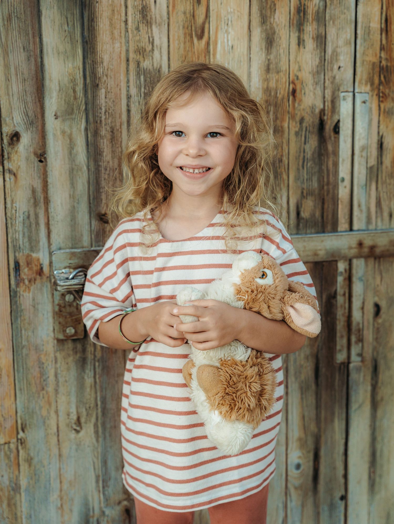 Smiling young girl in striped dress holds a stuffed animal against a rustic wooden door.