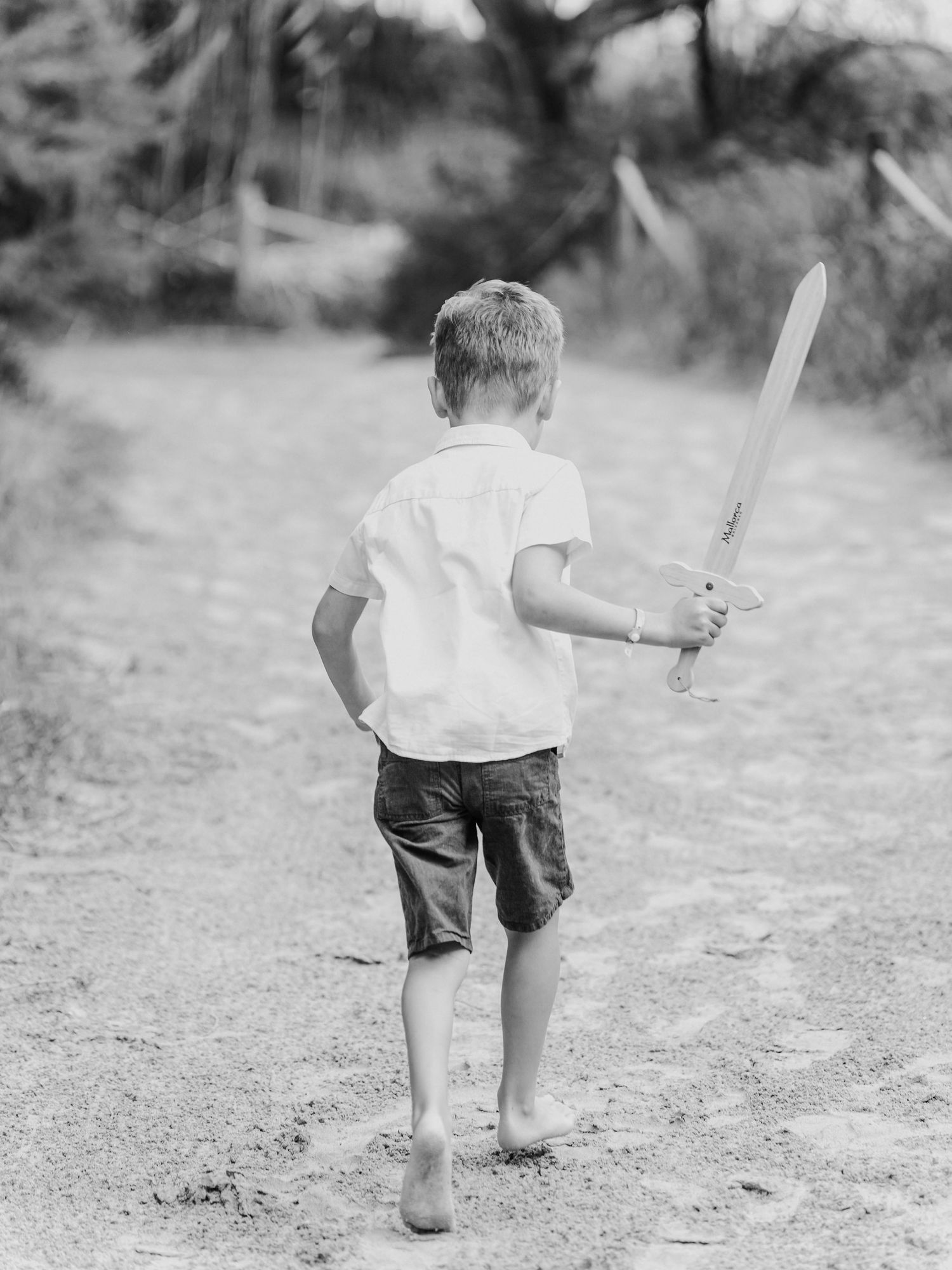 Black and white photo of a barefoot boy walking on a sandy trail holding a wooden toy sword.