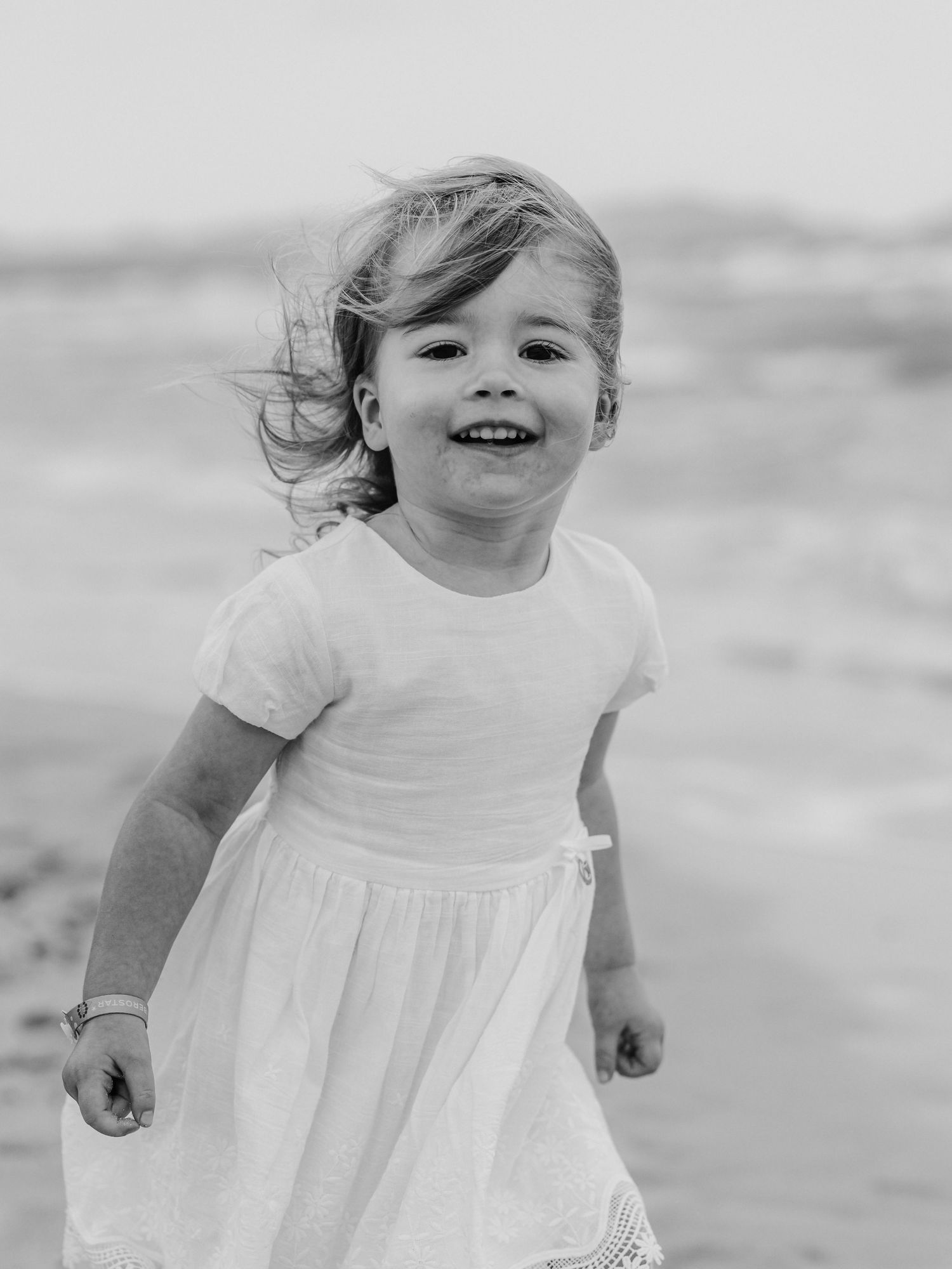 Black and white photo of a smiling little girl in white dress running on a beach with wind in her hair.