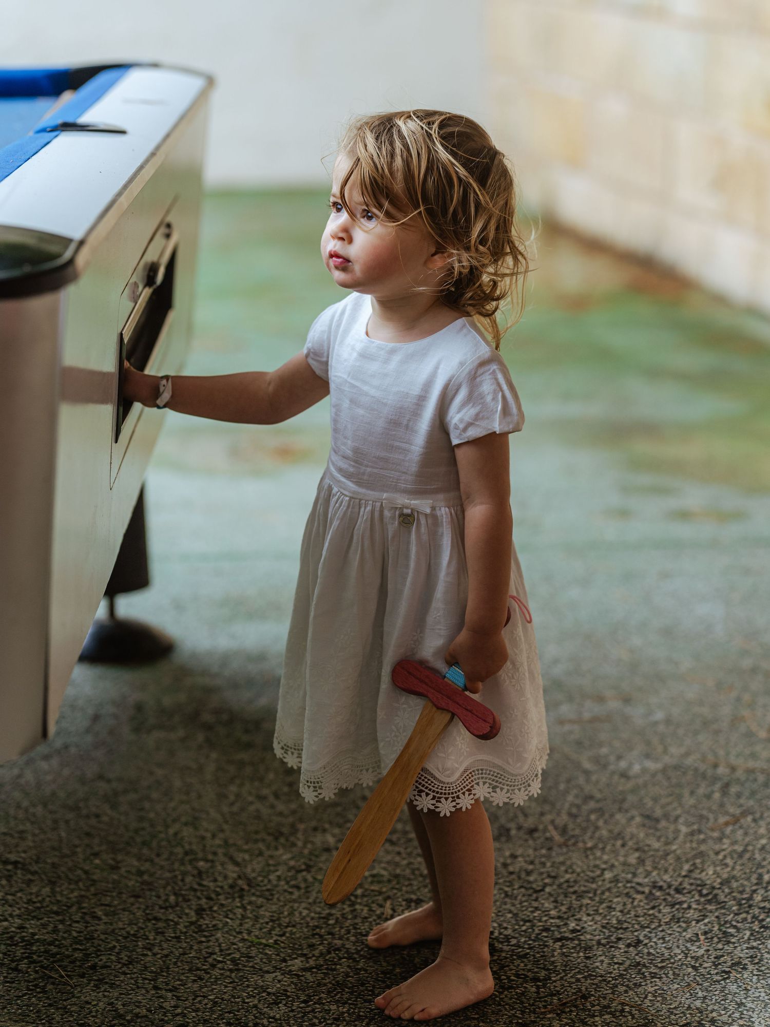 Barefoot toddler girl in white lace dress holding wooden toy while touching a white appliance.