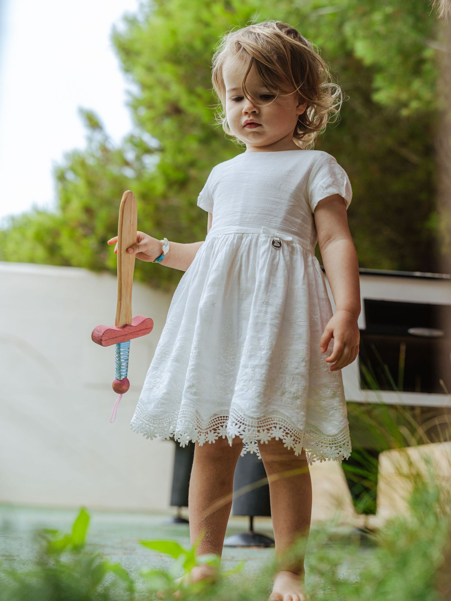 Barefoot toddler girl in white lace dress looking down at a wooden toy sword she is holding.